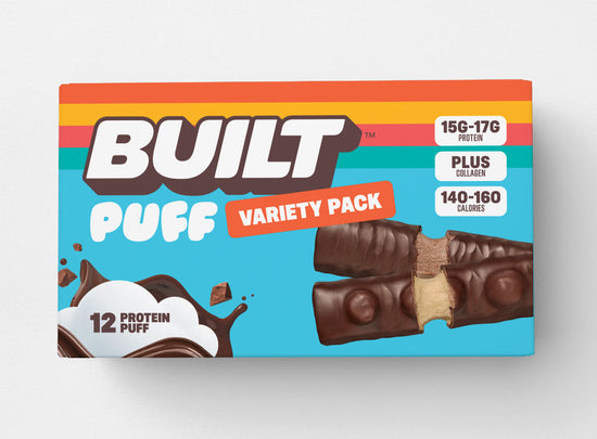 Stack of 12 BUILT mixed snack boxes with assorted flavors in a cardboard carton, viewed from the back.